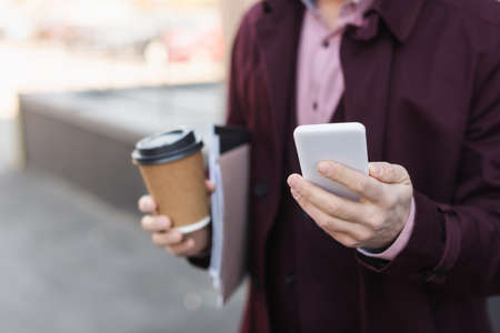 Cropped view of blurred businessman with paper cup and folders holding smartphone outdoorsの写真素材