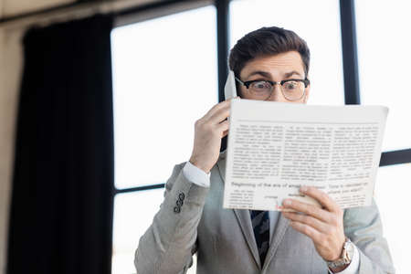 Shocked businessman talking on smartphone and looking at blurred newspaper in officeの写真素材