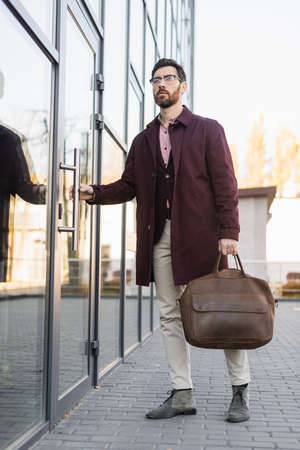 Businessman in trench coat holding bag near door of building outdoorsの写真素材