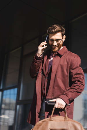 Smiling businessman in trench coat holding bag and notebook while talking on smartphone outdoorsの写真素材