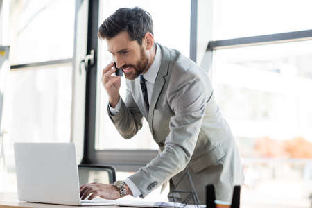 Positive businessman in formal wear talking on smartphone and using laptop in office