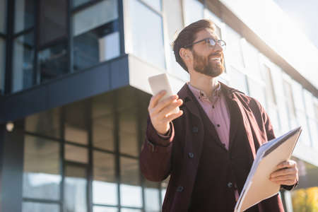 Low angle view of smiling businessman holding paper folder and using mobile phone outdoors