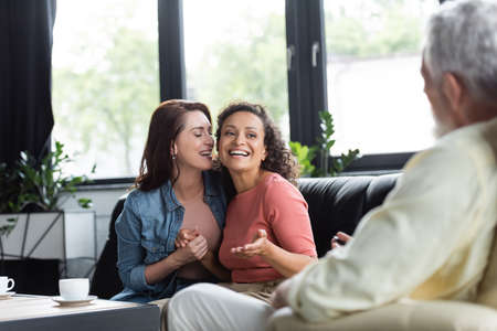 happy african american american woman talking to blurred psychologist while sitting on sofa with lesbian girlfriendの写真素材