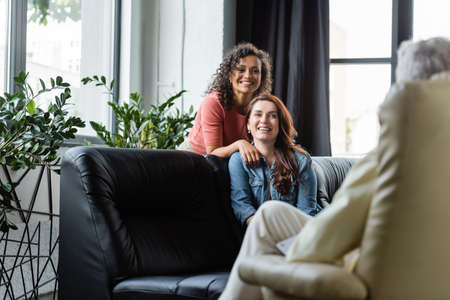 cheerful multiethnic lesbians looking at blurred psychologist during consultation