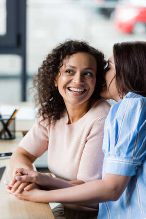 lesbian woman whispering in ear of cheerful african american girlfriend while sitting in real estate agencyの写真素材