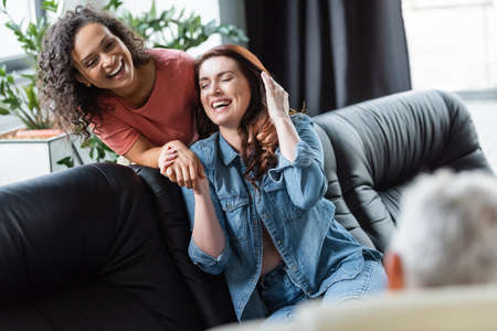 excited interracial lesbian couple holding hands and laughing near blurred psychologist during...