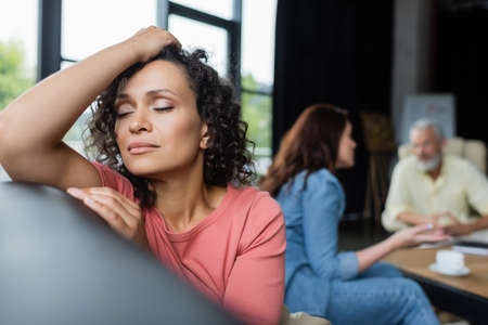 depressed african american woman sitting with closed eyes near girlfriend and psychologist on blurred backgroundの写真素材