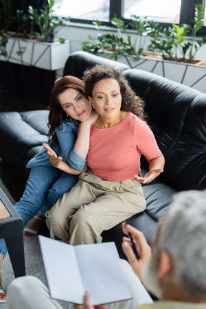 african american woman talking to blurred psychologist with notebook while sitting on sofa near lesbian girlfriendの写真素材