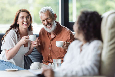 smiling couple sitting with coffee cups near blurred african american psychologistの写真素材