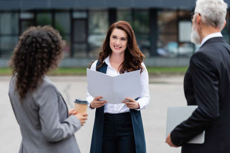 real estate agent with folder smiling near blurred multiethnic business partners outdoorsの写真素材
