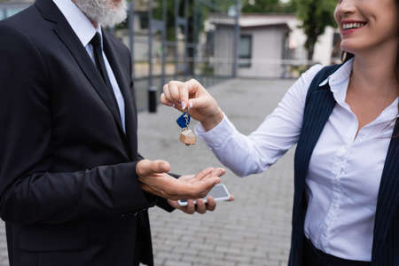cropped view of smiling realtor giving key to businessman holding smartphone outdoorsの写真素材