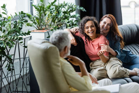cheerful multiethnic lesbians sitting on couch near blurred psychologist