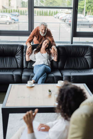happy man embracing smiling wife sitting on sofa in front of blurred african american psychologistの写真素材