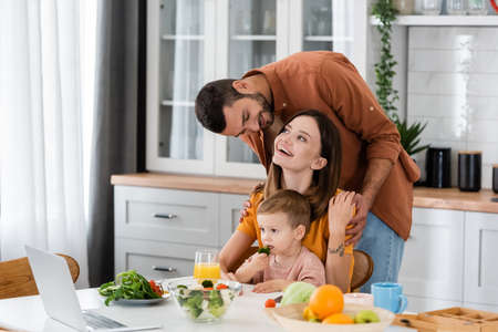 Positive man hugging wife near son, salad and laptop in kitchenの写真素材