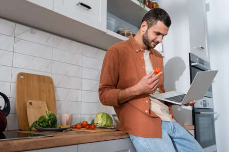 Young man holding cherry tomato and laptop near vegetables in kitchenの写真素材