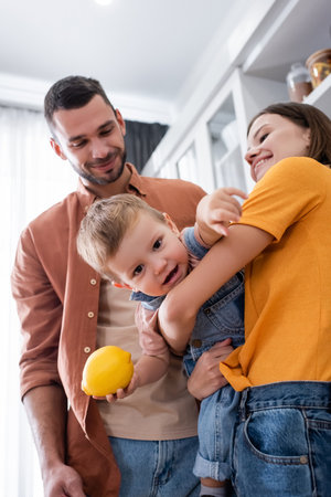 Smiling mother holding toddler son with lemon near husband at homeの写真素材