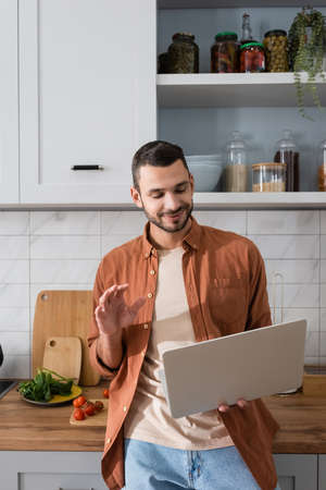 Smiling man having video call on laptop near vegetables in kitchen