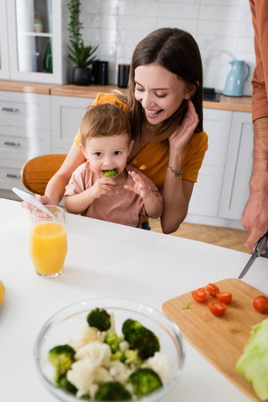 Happy mother holding smartphone and looking at child eating broccoli at homeの写真素材