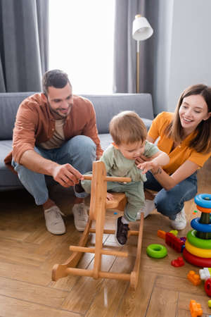 Happy parents holding kid near rocking horse and toys at homeの写真素材