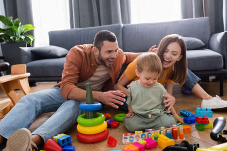 Positive parents hugging toddler kid near toys and bike in living roomの写真素材