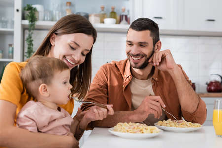 Smiling man looking at wife feeding son with pasta near orange juice in kitchenの写真素材