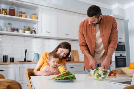 Happy woman holding orange juice near son and husband cutting vegetables in kitchenの写真素材