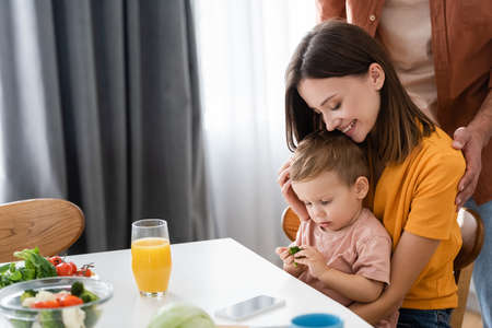 Cheerful woman hugging kid with broccoli near husband and salad at homeの写真素材