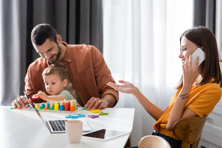 Smiling woman talking on cellphone near gadgets and blurred family at homeの写真素材