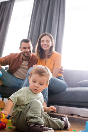 Toddler boy playing building blocks near blurred parents on couch in living roomの写真素材