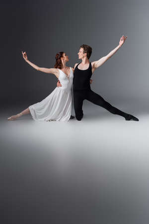 full length of graceful ballerina in white dress dancing with partner looking at each other on dark greyの写真素材