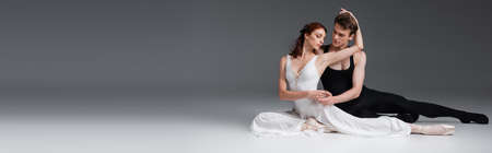full length of graceful ballerina in white dress sitting with partner on dark grey, bannerの写真素材