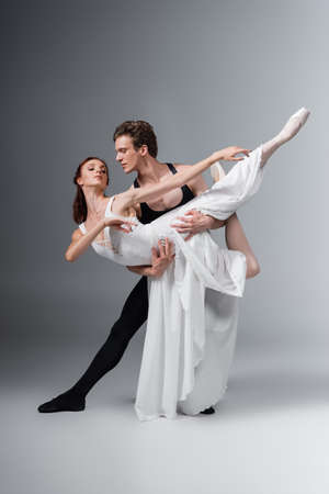 full length of man supporting graceful ballerina in white dress while dancing on dark greyの写真素材