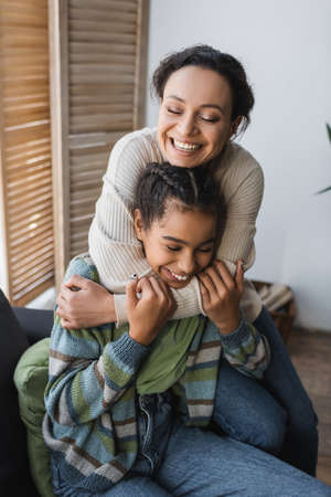 cheerful african american woman with teenage daughter embracing with closed eyes at homeの写真素材