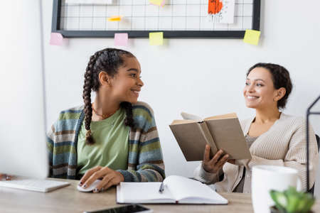 african american teenage girl smiling near computer and blurred mother with bookの写真素材
