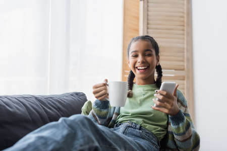 cheerful african american teen girl looking at camera while holding smartphone and cup of tea