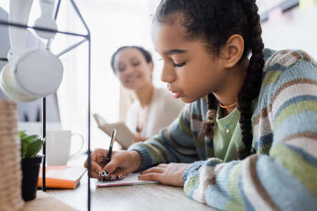 african american girl writing in notebook while doing homework near headphones and blurred momの写真素材