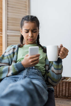 displeased african american teenage girl with cup of tea messaging at cellphone at home