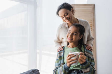 pleased african american woman hugging shoulders of teenage daughter holding tea cupの写真素材