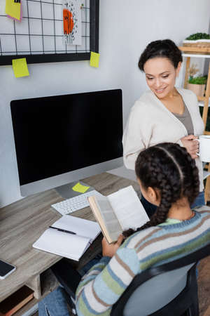 african american girl reading book near monitor with blank screen and smiling mom with cup of teaの写真素材