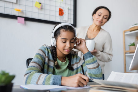 african american girl in headphones writing in notebook near blurred mom with cup of teaの写真素材