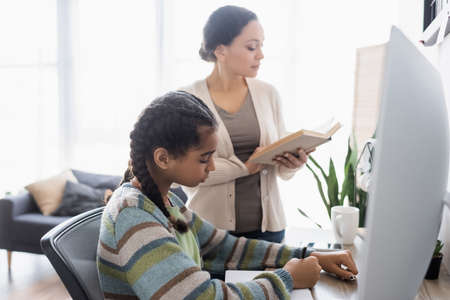 african american girl writing in notebook near blurred monitor and mother reading bookの写真素材