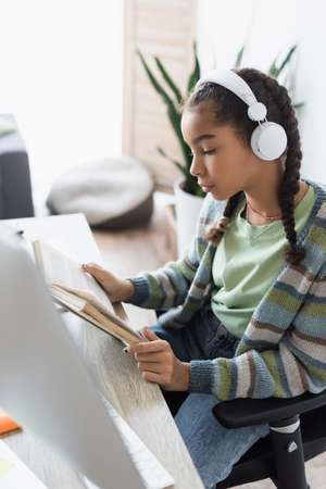 african american schoolgirl in headphones reading book near blurred computer monitor