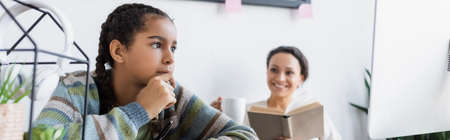 african american teen girl thinking near computer monitor and smiling mother with book on blurred background, bannerの写真素材