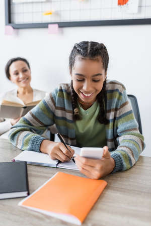 happy african american teenage girl writing in notebook near mother with book smiling on blurred backgroundの写真素材