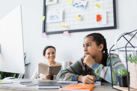thoughtful african american girl doing homework near laptop and mom with book smiling on blurred...