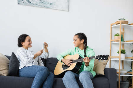 african american woman with smartphone taking photo of daughter playing guitar at home