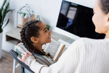 blurred african american woman standing near smiling daughter sitting with book near monitor with blank screenの写真素材