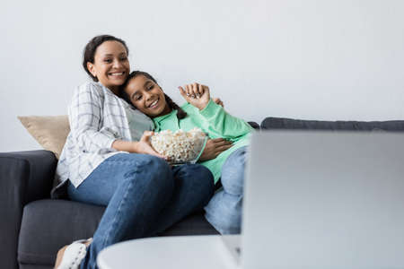 happy african american mom and daughter with bowl of popcorn watching movie on blurred laptop