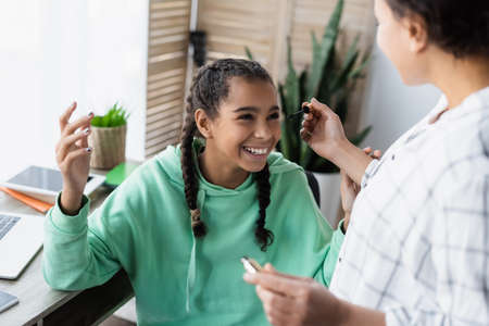 blurred african american woman holding mascara while doing mascara to joyful daughter