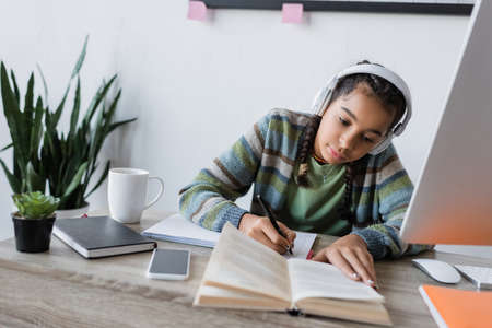 african american girl in headphones writing near book and computer while studying at homeの写真素材